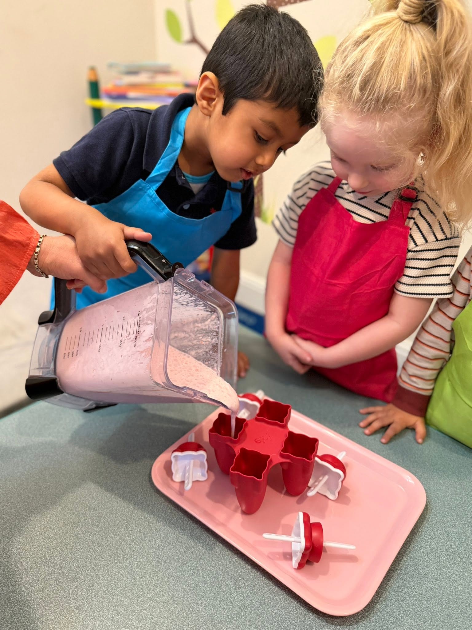 Two children in aprons pouring a strawberry smoothie into popsicle molds during a cooking project