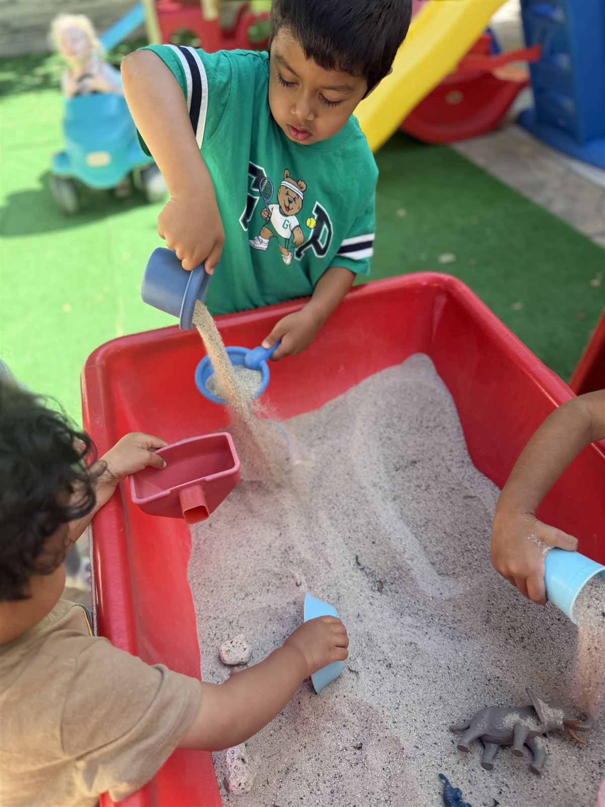 Children scooping and pouring sand together at an outdoor sand table beside the play structures
